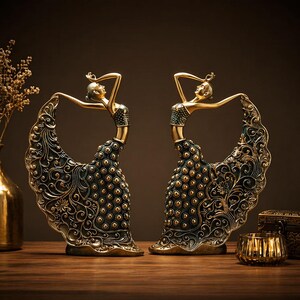 May include: Two gold-toned statues of dancers in flowing gowns. The figures have their arms raised, with intricate detailing on the dresses and bases. The dresses are black with gold accents and a pattern of small gold spheres.