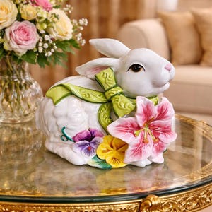 May include: A white ceramic rabbit ornament with a green bow and pink and yellow floral details. The rabbit has brown eyes and is on a glass table. A vase of flowers is in the background.