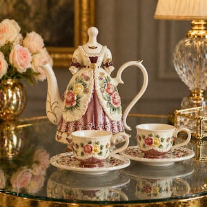 Könnte beinhalten: Dekoratives Porzellan-Teeservice mit einer Teekanne in Form eines Kleides, passenden Tassen und Untertassen. Das Set ist cremefarben mit floralen Designs in Rot, Gelb und Lila. Die Teekanne ist etwa 25 cm hoch.
