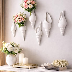 May include: White ceramic seashell-shaped wall vases, some filled with pink and white roses. A white vase with white flowers, candles, and a decorative tray sit on a marble-topped table. A coral decoration and books are also on the table.