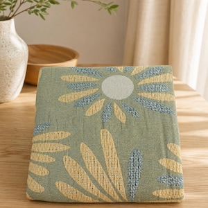 May include: A folded green textile with a large floral pattern in yellow and blue. The design features a central circle with radiating petals. The textile is placed on a wooden surface, with a vase and wooden bowl in the background.