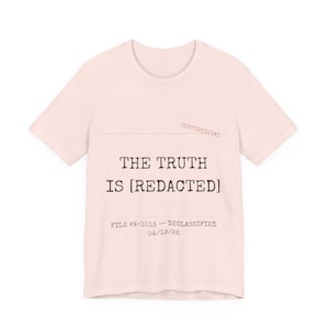 Può includere: T-shirt rosa chiaro con la scritta "THE TRUTH IS [REDACTED]" in nero, carattere di macchina da scrivere. La parola "CONFIDENTIAL" è stampata in rosso in alto a destra. Sotto, "FILE #X-1013 - DECLASSIFIED 04/19/26".