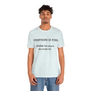 May include: Light blue t-shirt with the text "EVERYTHING IS FINE." and the source: "The people who broke it." The shirt is a crew neck and has short sleeves. The person in the image is wearing blue jeans.