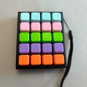 Può includere: Un colorato puzzle portatile con sedici blocchi quadrati in una griglia 4x4. I blocchi sono disposti in file di blu pastello, rosa, verde, viola e arancione. Il puzzle è inserito in una cornice nera e ha un cordino nero attaccato.