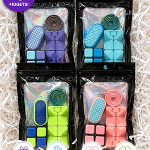 Può includere: Quattro set di giocattoli sensoriali fidget confezionati in buste olografiche nere individuali. Ogni set include una bobina circolare, un ovale testurizzato, un blocco multi-cubo e una piccola griglia quadrata. Testo sull'immagine: INCLUDES 4 FIDGETS!