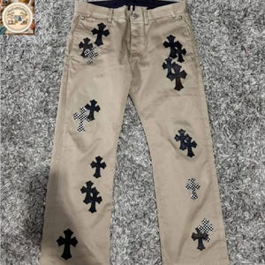 May include: Khaki trousers featuring multiple black cross appliqués. Some crosses have a chequered pattern. The trousers have a classic five-pocket design and a button and zip closure. The trousers are made of a durable material.