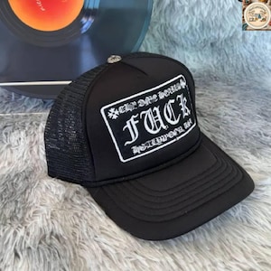 Puede incluir: Gorra de camionero negra con parte trasera de malla y visera curva. La parte delantera presenta un parche rectangular blanco con la palabra "FUCK" en letras góticas y el texto "Chrome Hearts Hollywood, CA".