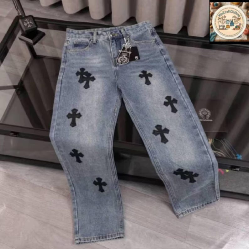 May include: Light blue denim jeans featuring multiple black cross appliques. The jeans have a classic five-pocket design and a straight leg fit. A metal tag hangs from the belt loop. The jeans are displayed on a reflective surface.