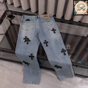 May include: Light blue denim jeans with black cross appliques. The jeans have a classic five-pocket design, a button and zipper closure, and a leather patch on the back. The crosses are placed on the front and back of the jeans.