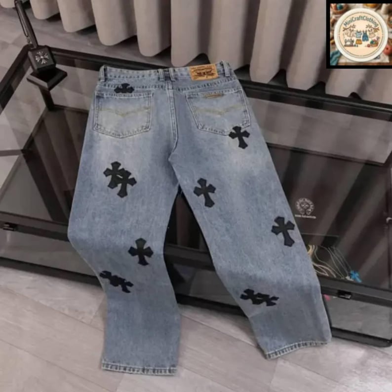 May include: Light blue denim jeans with black cross appliques. The jeans have a classic five-pocket design, a button and zipper closure, and a leather patch on the back. The crosses are placed on the front and back of the jeans.