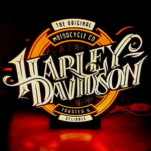 Può includere: Un'insegna luminosa circolare che presenta il testo Harley Davidson in un carattere bianco stilizzato. Lo sfondo è un cerchio arancione brillante con il testo The Original Motorcycle Co in alto e Trusted & Reliable in basso.