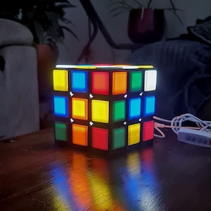 Op de afbeelding: Een vierkante, meerkleurige LED-lamp in de vorm van een puzzelkubus. De lamp heeft een zwart rasterframe met verlichte panelen in geel, blauw, rood, oranje, groen en wit. Een wit netsnoer met een klein bedieningspaneel is aan de zijkant bevestigd.