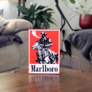 Può includere: Un'insegna rettangolare con sfondo rosso e un'illustrazione grafica in bianco e nero di un cowboy a cavallo. La parola Marlboro è stampata in lettere maiuscole nere nella parte inferiore dell'insegna su un bordo rettangolare bianco.