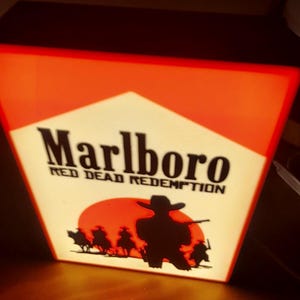 Può includere: Un'insegna luminosa rettangolare con un design rosso e bianco e la scritta Marlboro e Red Dead Redemption in nero. La grafica include la sagoma nera di un cowboy con cappello davanti a un sole rosso e alcune figure a cavallo.