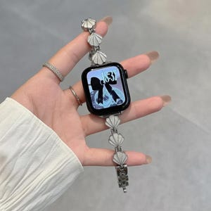 May include: A smartwatch with a black rectangular face displaying a digital clock showing 18:37 and a black bow graphic. The watch band is made of silver-toned metal links, each featuring a small, white, fan-shaped shell design element.