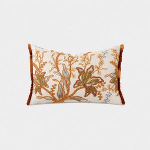 May include: A rectangular throw cushion featuring an off-white base with intricate embroidery in shades of burnt orange, olive green, and muted blue. The design depicts stylised floral and botanical motifs, finished with a textured fringe trim.