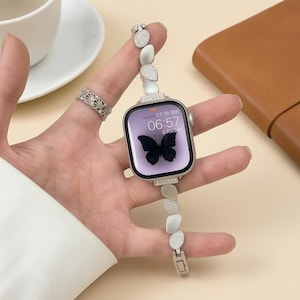 Op de afbeelding: Een zilverkleurige smartwatch met een rechthoekig scherm waarop een zwarte vlinder en de tijd 06:57 worden weergegeven. De horlogeband heeft afwisselend gladde zilverkleurige ovale schakels en schakels met kristallen. Een bijpassende ring.