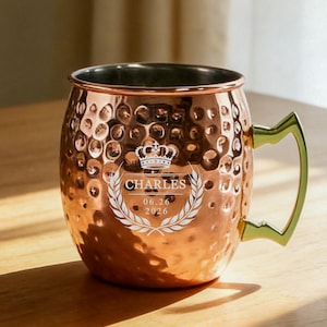 Pode incluir: Uma caneca martelada de cor cobre com uma alça de metal dourado. A frente apresenta um design gravado em branco de uma coroa acima do nome CHARLES, a data 06.26.2026 e uma moldura de louros. O interior da caneca é prateado.