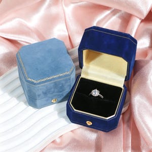 Könnte beinhalten: Zwei Samt-Ringboxen, eine geschlossene und eine offene, die einen Diamantring zeigen. Die offene Box ist marineblau mit goldener Innenausstattung, die geschlossene Box ist hellblau. Beide haben goldene Verzierungen und Verschlüsse, vor rosa Satin.