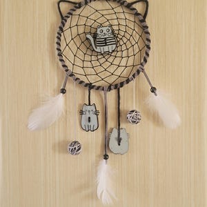 Op de afbeelding: Een dromenvanger met kattenmotief met een zwart web en grijze accenten. De dromenvanger heeft een katten silhouet in het midden, met hangende katten uitsparingen, witte veren en decoratieve ballen. Het ontwerp is geplaatst tegen een lichtgekleurde achtergrond.