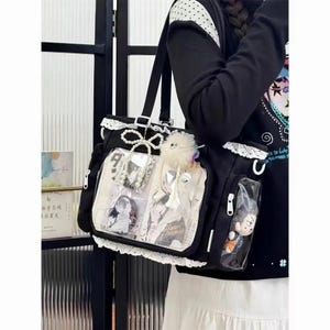 Könnte beinhalten: Schwarze Tragetasche mit weißer Spitzenborte und einer transparenten Vordertasche mit Anime-Artwork. Die Tasche hat eine Schleife und einen flauschigen Anhänger. Zusätzliche transparente Taschen halten kleine Figuren.