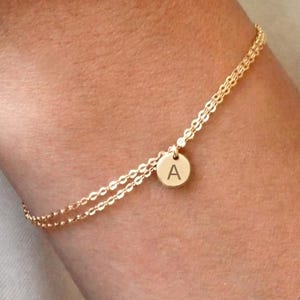 Könnte beinhalten: Ein zartes, goldfarbenes Armband mit doppelter Kette und einem kleinen, runden Anhänger, in den der Großbuchstabe A eingraviert ist. Das Armband besteht aus zwei dünnen, miteinander verbundenen Metallketten am Handgelenk.
