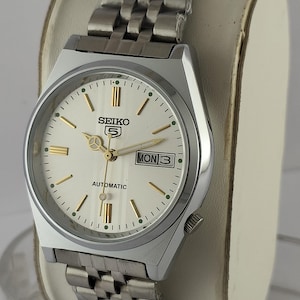 Op de afbeelding: Een Seiko 5 automatisch polshorloge met een zilverkleurige roestvrijstalen schakelarmband en een ronde kast. Het horloge heeft een crèmekleurige wijzerplaat met goudkleurige uurmarkeringen en wijzers, een dag-datumvenster op drie uur en tekst.