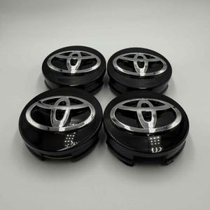 Könnte beinhalten: Vier schwarze Radnabenabdeckungen mit einem silbernen Toyota-Logo. Die Kappen sind rund mit glänzendem Finish. Das Toyota-Emblem ist ein Oval mit drei sich überlappenden Bögen.