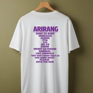 May include: White t-shirt with the word "ARIRANG" in purple block letters at the top, followed by a list of song titles in purple. The shirt is hanging on a wooden clothes hanger.