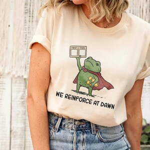 May include: A cream-colored t-shirt featuring a graphic of a green frog wearing a red cape and holding a sign that reads FIRST THEN. The frog is also holding several yellow stars. Below the graphic, the text reads WE REINFORCE AT DAWN.