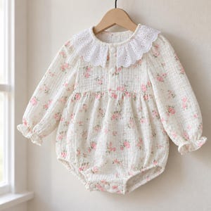 May include: A long-sleeved romper in a cream-coloured fabric with a small pink floral print. The garment features a white eyelet lace collar, a button-front placket with two white buttons, and elasticated cuffs. The bottom includes three snap closures.