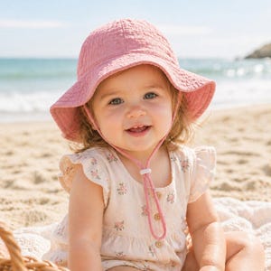 Könnte beinhalten: Ein Baby trägt einen rosa Sonnenhut mit Kinnriemen und ein Kleid mit Blumenmuster an einem Sandstrand. Der Hut hat eine breite Krempe und ein strukturiertes Aussehen. Das Kleid hat einen hellen Hintergrund mit rosa Blumenmustern.