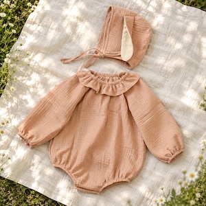May include: A dusty rose long-sleeve romper featuring a ruffled collar and elasticated cuffs, paired with a matching bonnet that includes two long, pointed fabric ears and a tie closure under the chin, all crafted from a textured muslin fabric.