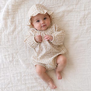 May include: A baby wearing a long-sleeved beige and white gingham romper with elasticated cuffs and a matching ruffled bonnet. The outfit features a gathered neckline and a soft, lightweight fabric construction, providing a coordinated look.