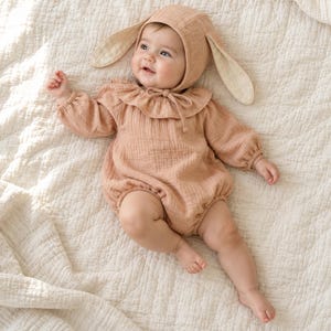 May include: A light peach-coloured long-sleeved romper with a ruffled collar and a matching bonnet featuring long, floppy bunny ears. The garment is made of a textured fabric and includes a tie closure at the neck, providing a soft, cohesive look.