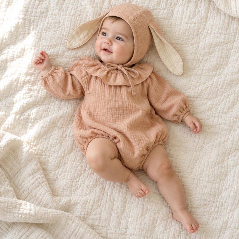 May include: A light peach-coloured long-sleeved romper with a ruffled collar and a matching bonnet featuring long, floppy bunny ears. The garment is made of a textured fabric and includes a tie closure at the neck, providing a soft, cohesive look.