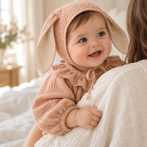 May include: A dusty rose-coloured long-sleeved top with a ruffled collar and a matching bonnet featuring long, floppy rabbit ears. The bonnet is tied under the chin with thin fabric strings, and the garment is made of a textured, crinkled fabric.