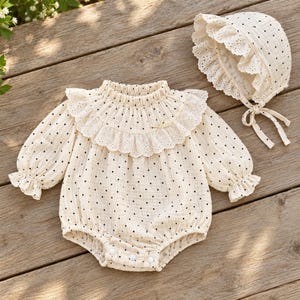 May include: A cream-coloured long-sleeve romper featuring a black polka dot pattern, a ruffled eyelet collar with a small bow, and elasticated cuffs. The set includes a matching bonnet with a ruffled eyelet trim and tie closures under the chin.
