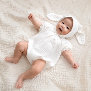 May include: A white muslin baby romper with short puff sleeves and a matching bonnet featuring long, floppy rabbit-style ears. The garment is crafted from a lightweight, textured fabric and is designed with a gathered neckline and elasticised leg openings.