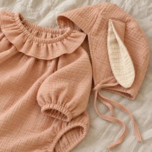 May include: A dusty rose-coloured long-sleeve romper featuring a ruffled collar and gathered cuffs, paired with a matching bonnet that includes two long fabric ties and a single prominent rabbit-style ear detail in a lighter cream fabric.