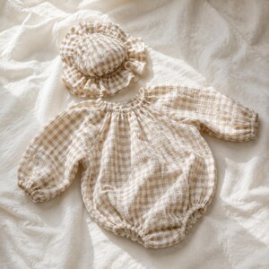 May include: A long-sleeved romper and a matching ruffled bonnet, both featuring a beige and white gingham check pattern. The romper has a gathered neckline and elasticised cuffs, while the bonnet has a circular crown and a wide, ruffled brim.