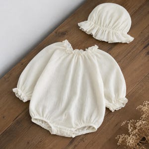May include: A cream-colored long-sleeve romper featuring a ruffled neckline and elasticized ruffled cuffs, paired with a matching cream-colored bonnet that has a wide, gathered ruffled brim. The fabric has a subtle, crinkled, textured appearance.