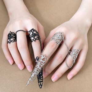 May include: Two-finger rings with intricate, openwork designs. One ring is black, the other is silver-toned, connected by delicate chains. The rings feature floral and geometric patterns, creating a gothic or fantasy aesthetic.
