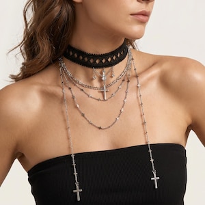 May include: A black lace choker necklace with a diamond pattern, layered with multiple silver necklaces. The necklaces feature cross and star pendants, creating a gothic or religious aesthetic. The jewelry is worn against a black strapless top.