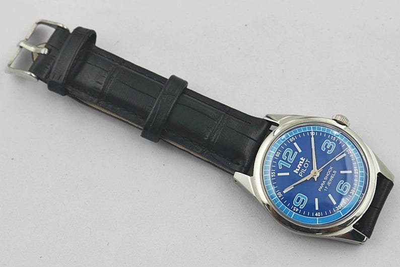 May include: A wristwatch with a blue face and a black leather band. The watch face has white numerals and the text "hmt PILOT PARA SHOCK 17 JEWELS". The band has a silver buckle.