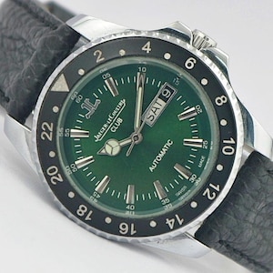 May include: A Jaeger-LeCoultre Club automatic wristwatch with a green dial, silver-tone hands and hour markers, and a black 24-hour rotating bezel. The watch features a day-date window at the three o'clock position and a black textured leather strap.