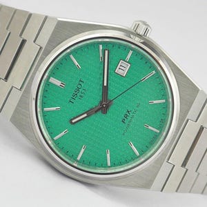 Puede incluir: Un reloj Tissot PRX Powermatic 80 con brazalete y caja integrados de acero inoxidable cepillado. La esfera es de un verde vibrante con textura de gofre, índices de bastón plateados, manecillas luminiscentes y una ventana de fecha a las tres.