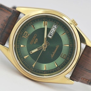 May include: A gold-tone Seiko 5 automatic watch with a round green sunray dial, gold-tone hands and indices, and a brown leather strap with a textured finish. The dial features the text Seiko 5, Automatic, and a day-date window displaying WED 22.