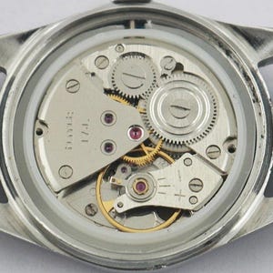 May include: Close-up of a watch's inner workings, showcasing intricate gears, jewels, and a silver-toned metal frame. The watch has a black leather strap. The mechanical components are visible through the clear back.