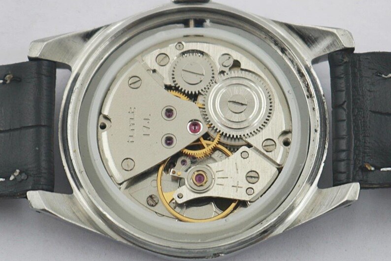 May include: Close-up of a watch's inner workings, showcasing intricate gears, jewels, and a silver-toned metal frame. The watch has a black leather strap. The mechanical components are visible through the clear back.
